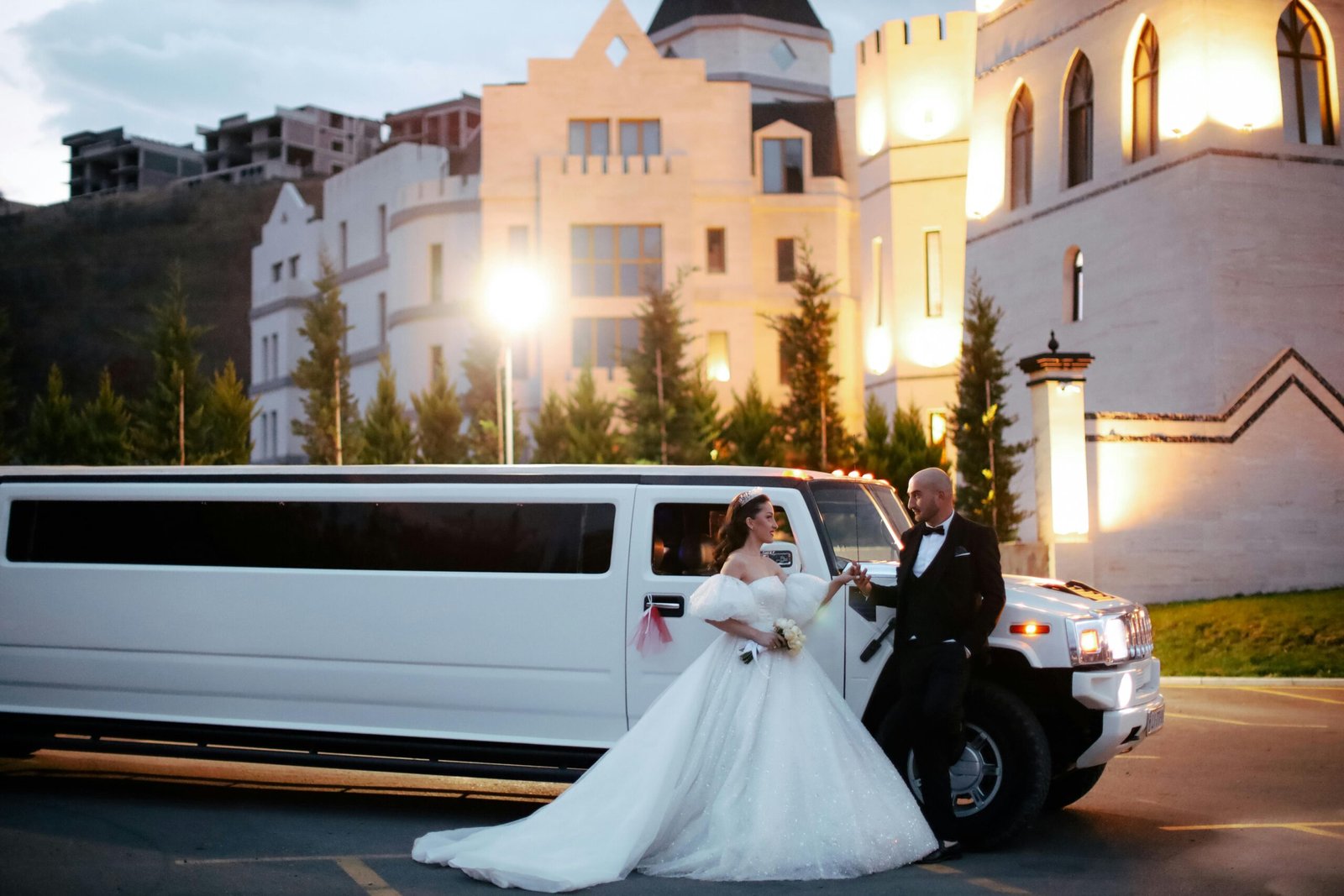 Bride and groom in formal attire with a limousine in front of an illuminated building at night.